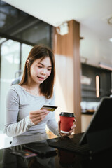 Fototapeta premium A young woman holding a credit card and coffee cup shops online using a laptop at a modern café, looking focused and happy.