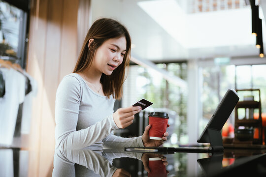 A young woman holding a credit card and coffee cup shops online using a laptop at a modern café, looking focused and happy.