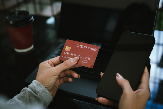 A young woman holding a credit card and coffee cup shops online using a laptop at a modern café, looking focused and happy.