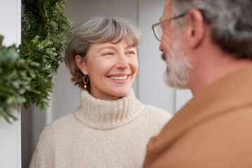 Happy senior couple together at home entrance during winter. smiling mature woman with gray hair looks lovingly at man while standing by festive wreath