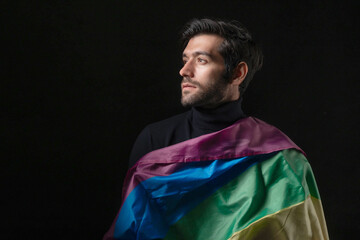 Confident young man wrapped in rainbow pride flag looking away with thoughtful expression, portrait of male supporting LGBTQ community, diversity, equality, and freedom of identity on dark background