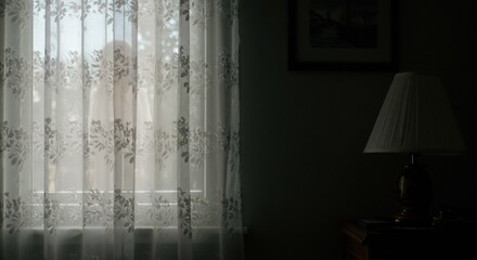 A shadowed room featuring a window veiled by a floral patterned curtain and a lit lamp on a bedside table