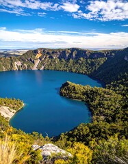 High-angle view of a vibrant blue crater lake, surrounded by lush green hills under a partly cloudy sky