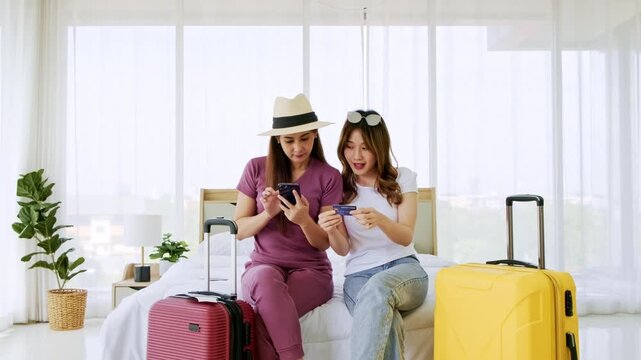 Two cheerful travelers sit on a hotel bed with suitcases, celebrating a booking while looking at a smartphone and sipping drinks in a bright room with large windows.