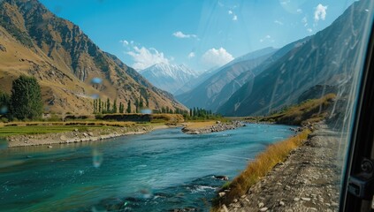A scenic view of a turquoise river winding through a mountainous valley on a bright sunny day, possibly seen from inside a vehicle.  The mountains are rugged and brown with patches of green vegetation