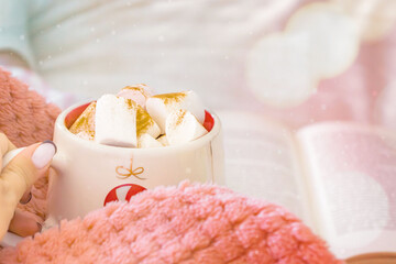 Close-up of a festive ceramic mug filled with hot chocolate topped with marshmallows and cinnamon powder. On the background is an open book and a bowl with colorful candy. Concept of comfort, hygge