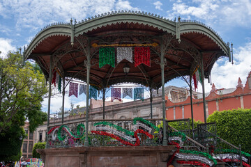 Obraz premium A colorful, multi-colored gazebo with flags hanging from it. The gazebo is surrounded by trees. Historic center of Queretaro City, decorations and traditions to celebrate Mexico's Independence Day, co