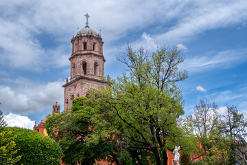 A tall tower with a cross on top stands in front of a green tree. The sky is blue. Historic center...