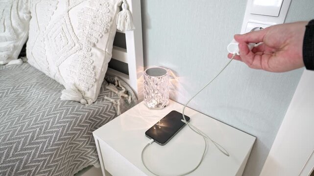 Plugging smartphone charger into wall socket near bed and night light, then unplugging in bright, minimalist bedroom setting with warm, clean ambiance
