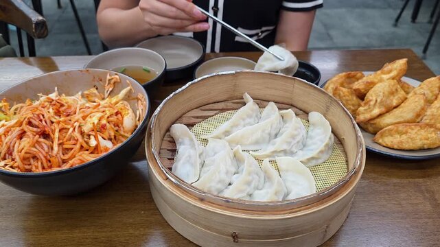 A woman uses chopsticks to pick up a delicious steamed mandu from a bamboo steamer, part of a traditional Korean meal that also includes fried dumplings and spicy noodles at an authentic restaurant