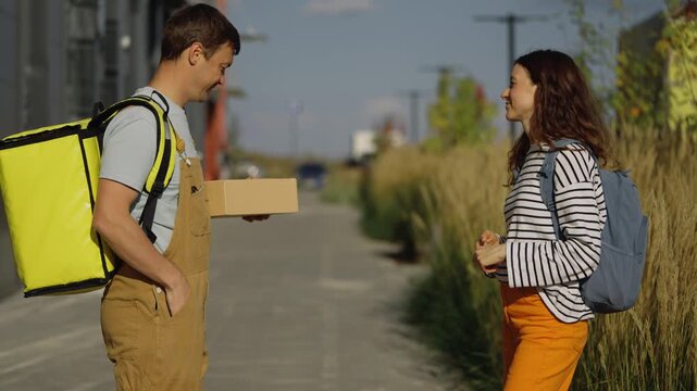 Young woman receiving a small cardboard box from a friendly delivery man wearing a thermal backpack, confirming the order on his smartphone before leaving on a sunny city street
