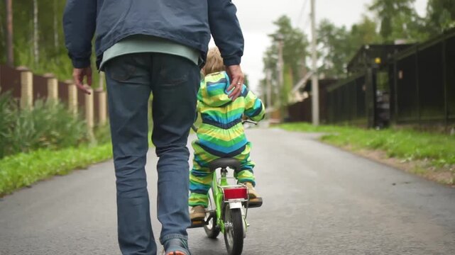 Dad supporting young child learning to ride bicycle without helmet, demonstrating importance of safety gear during family sports activity.