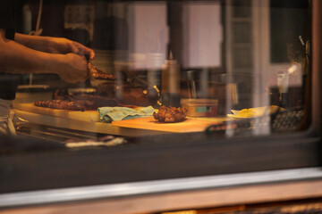 Viewed from outside glass show window, Japanese chef prepares unagi or freshwater eel meal on wooden counter next to waiting teriyaki chicken breast. Chef's hands are busy skewering grilled unagi.