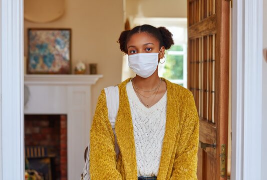 A young woman with pigtails, wearing a medical mask and a yellow sweater, stands at an open doorway, looking directly at the camera - Powered by Adobe