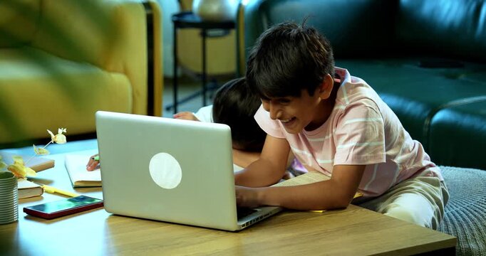 Indian small brother sister studying at home with laptop textbook, notepad indoors, Asian young kids siblings learning together using computer technology in modern living room, education and homework