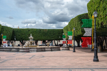 A park with a fountain and trees with lights on them. The lights are red, white, and green. Historic center of Queretaro City, decorations and traditions to celebrate Mexico's Independence Day, coloni