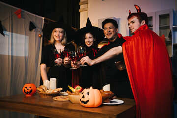 A cheerful group of friends dressed as witches and devils celebrate Halloween together,raising glasses of wine around a festive table with pumpkins,candles, bread,decorations in  cozy indoor setting
