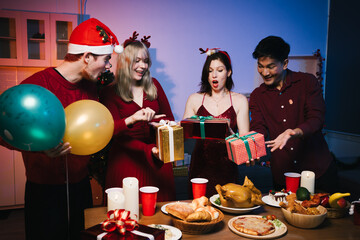 Four joyful friends gather for a festive Christmas party, exchanging colorful wrapped gifts around a table filled with delicious holiday food, balloons, and candles, creating a warm, cheerful