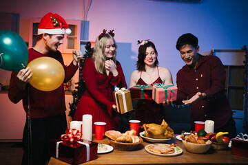 Four joyful friends gather for a festive Christmas party, exchanging colorful wrapped gifts around a table filled with delicious holiday food, balloons, and candles, creating a warm, cheerful