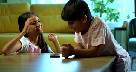 Indian small brother sister playing mobile games on smartphone devices spending leisure time at home, Asian young kids siblings enjoying fun childhood activity together indoors in modern living room