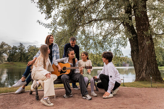 multigenerational group of friends plays guitar and sings by the lake.