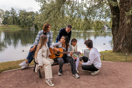 multigenerational group of friends plays guitar and sings by the lake.