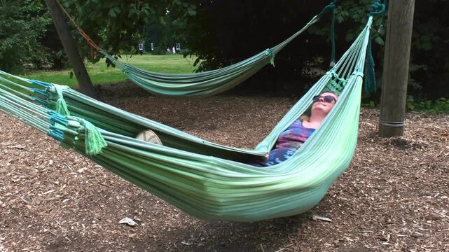 A woman gently sways in a hammock between trees in a sunlit park, enjoying a peaceful moment surrounded by greenery and calm nature. - Powered by Adobe