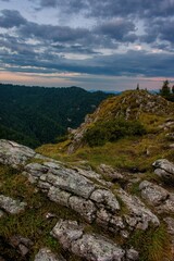 Rocky mountain summit with grassy slopes and cloudy sunset sky, overlooking a vast forested valley. Scenic view from a hiking trail in the wilderness.