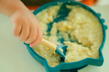 Detailed shot of toddlers hand holding spoon in bowl of healthy porridge, showing self-feeding during weaning and independent eating development.