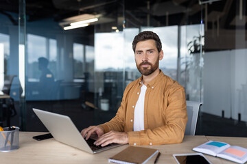 Young businessman sitting at a wooden desk, confidently looking at the camera while typing on a laptop, surrounded by modern office technology and a glass-paneled workspace
