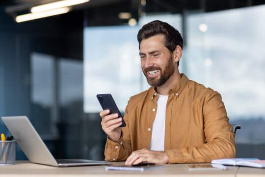 Happy businessman smiling at his desk while using a smartphone and laptop, connecting online for work-chatting, emailing and video calling in a modern office environment - Powered by Adobe