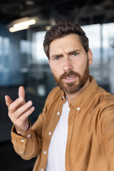 Confident businessman looking at webcam, actively talking, explaining, and gesturing during a virtual conference or online meeting, communicating remotely from a modern office