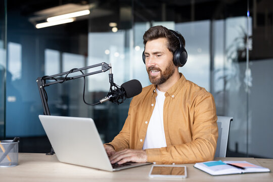 Man wearing headphones and speaking into a professional microphone while typing on a laptop, creating content for a podcast or live streaming online from a corporate workspace