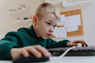 A boy with hearing aids focuses intently while playing a game on his laptop, showcasing modern...
