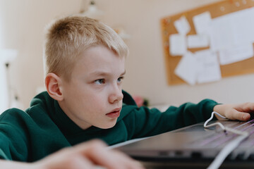 A boy with hearing aids focuses intently while playing a game on his laptop, showcasing modern...