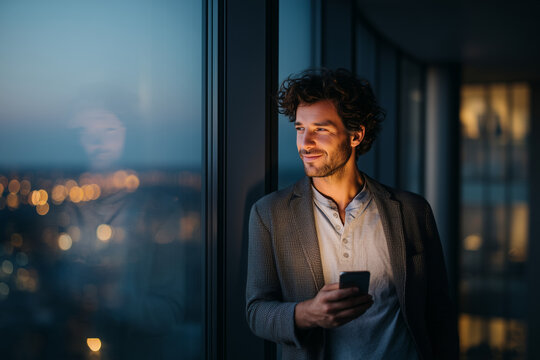Contemplative young man with smartphone gazing out of a high-rise window at city lights — warm evening glow, reflection and modern urban lifestyle portrait