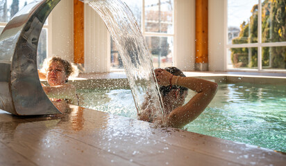 Two middle-aged female friends enjoying water massage from indoor pool waterfall at a spa center, representing relaxation, wellness and healthy lifestyle