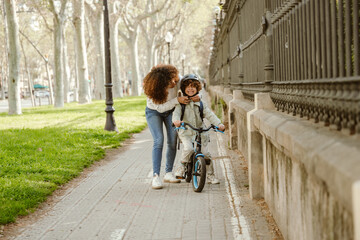 A mother stands and fastens a helmet on the head of her son who is sitting on a bicycle