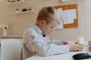 A boy with hearing aids in a button-down shirt enjoys a crisp apple at the kitchen counter. A natural moment of healthy snacking and inclusive childhood authenticity