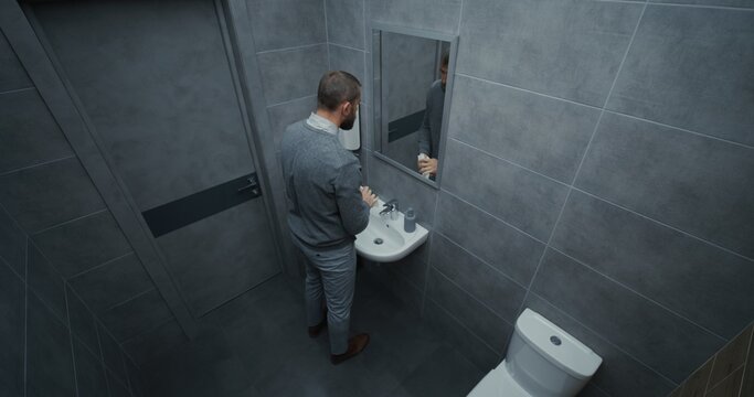 Businessman Looking in the Mirror, Washing His Hands with Soap in the Sink. Hand Hygiene after Using the Toilet. Man Doing Daily Routine in Restroom of Modern Office During Work. Lifestyle. High Angle - Powered by Adobe