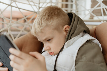 A boy with hearing aids in a jacket plays intently with a tablet on a cozy porch. Captures modern childhood, adaptive technology, and inclusive digital play in a natural setting © looking2thesky