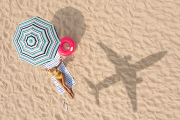 Woman resting under striped umbrella on beach, top view. Flying airplane casting shadow on sand