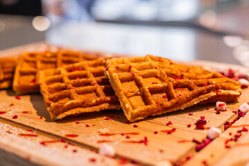 Golden waffles arranged on a wooden board, garnished with red sprinkles. The scene highlights a delicious breakfast or dessert option.