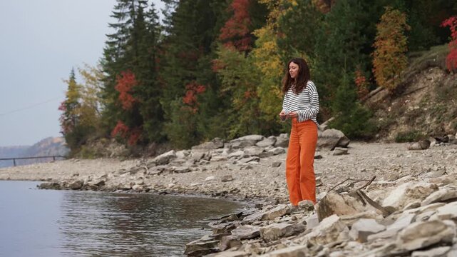 Young woman on a rocky lakeshore enjoying a quiet autumn day, skipping stones across the calm water surface amidst a beautiful forest with colorful foliage, finding a moment of peace