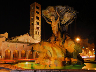 Beautiful baroque Fountain of the Tritons with medieval Basilica of Saint Mary in Cosmedin lit up at night in Rome