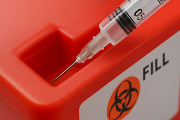 A close-up photograph showing a used syringe being properly disposed of in a red sharps container. The biohazard label emphasizes safe handling, infection control, and medical waste management in heal