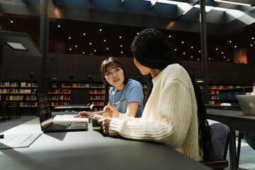 A female student listens and looks at a female student sitting next to her at a table with a laptop...