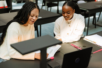 Two female students smiling and sitting at a table while one of them writes in a notebook