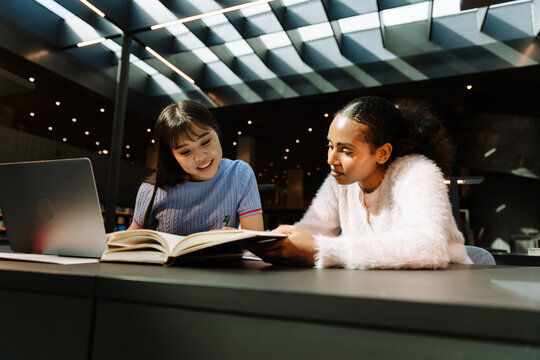 Two female students sitting at a table reading a book while one of them smiles