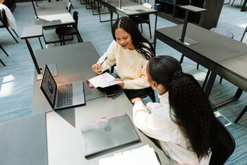 A female student is laughing and holding a pen and a notebook while sitting at a table with a female student